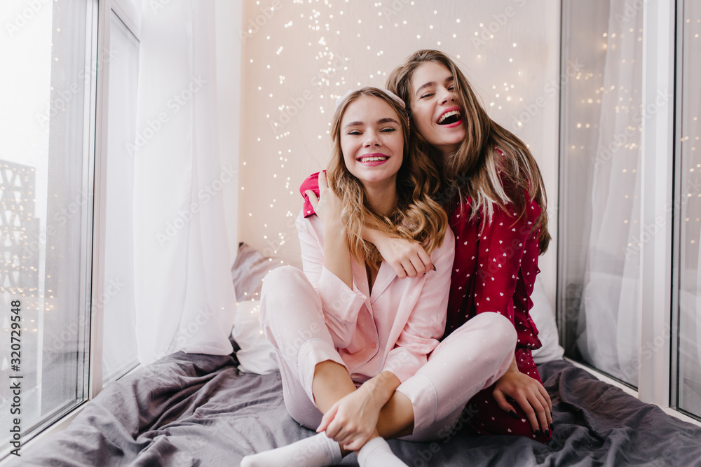 Obraz premium Cute european girl in pink sleepwear posing with charming smile in bedroom. Indoor portrait of fascinating ladies in pajamas hugging and laughing.