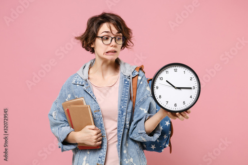 Confused young woman student in denim clothes eyeglasses, backpack posing isolated on pastel pink background. Education in high school university college concept. Mock up copy space. Hold books clock. photo