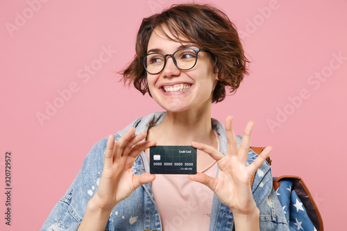 Smiling young woman student in denim clothes eyeglasses backpack posing isolated on pastel pink background. Education in high school university college concept. Looking aside, hold credit bank card.