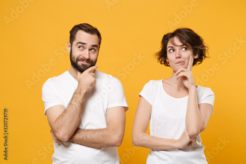 Pensive young couple friends bearded guy girl in white blank empty t-shirts posing isolated on yellow orange wall background. People lifestyle concept. Mock up copy space. Put hands prop up on chin.
