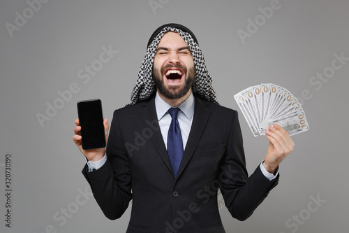 Laughing arabian muslim businessman in keffiyeh kafiya ring igal agal suit isolated on gray background. Achievement career wealth business. Hold mobile phone with blank empty screen fan of cash money. photo