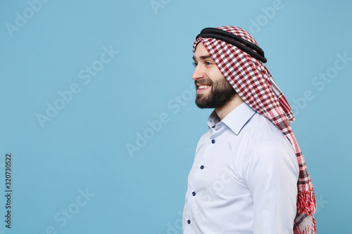 Photography Side view of smiling bearded young arabian muslim man in keffiyeh kafiya ring igal agal casual clothes posing isolated on pastel blue background