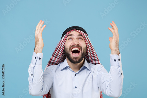 Crazy bearded arabian muslim man in keffiyeh kafiya ring igal agal casual clothes isolated on pastel blue background. People religious lifestyle concept. Mock up copy space. Screaming spreading hands. photo