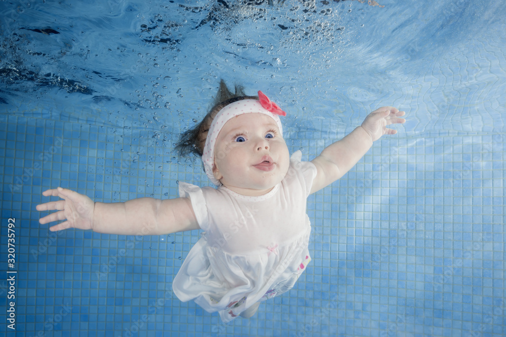 Little baby girl in white dress dives underwater in a swimming pool ...