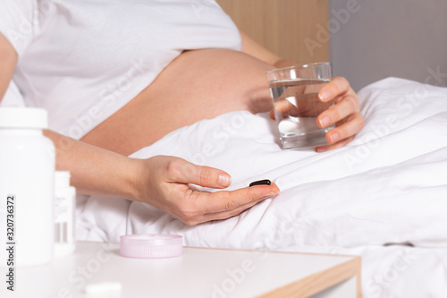 Unrecognizable Pregnant Woman in bed holding vitamins, pills in hand before intake. Glass of water is in another hand. Pregnancy healthy lifestyle. Snow-white bedding.