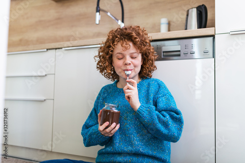 Cute ginger young woman in modern stylish clothes enjoying tasty chocolate spread with cute smile in the cuisine interier. young woman eating chocolate from a jar while sitting on the kitchen floor.