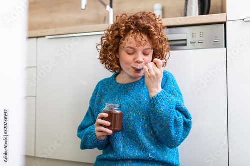 young woman eating chocolate from a jar while sitting on the wooden kitchen floor. Cute ginger girl indulging cheeky face eating chocolate spread from jar using spoon savoring every mouthful