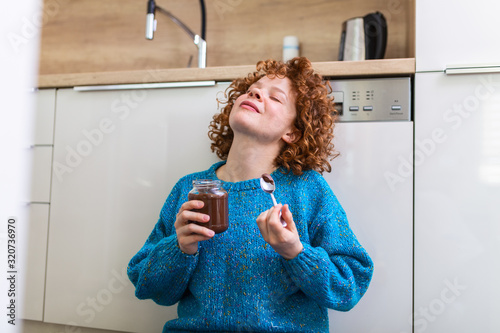 young woman eating chocolate from a jar while sitting on the wooden kitchen floor. Cute ginger girl indulging cheeky face eating chocolate spread from jar using spoon savoring every mouthful
