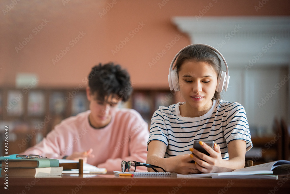 Pretty college student with headphones scrolling in smartphone while sitting by desk in reading hall of college library Pretty college student with headphones scrolling in smartphone while sitting by desk in reading hall of college library