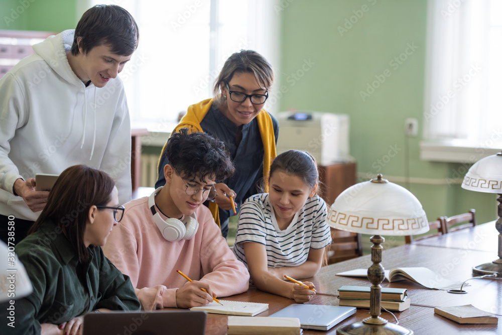 Obraz premium Two students pointing at notes of teenage guy in his notepad while choosing name for project in group in college library