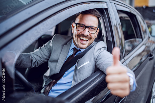 Handsome caucasian unshaven classy businessman in suit and with eyeglasses holding hand on steering wheel and giving thumbs up.