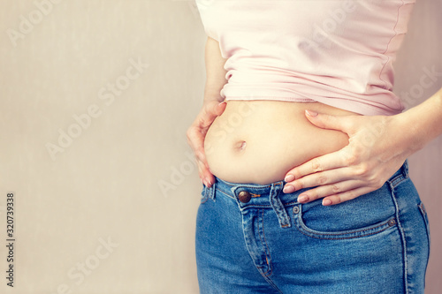 Woman in jeans and a light shirt is standing sideways and holding hands squeezes belly fat on light background, copy space. The concept of overweight, weight loss, diet, obesity, junk food.