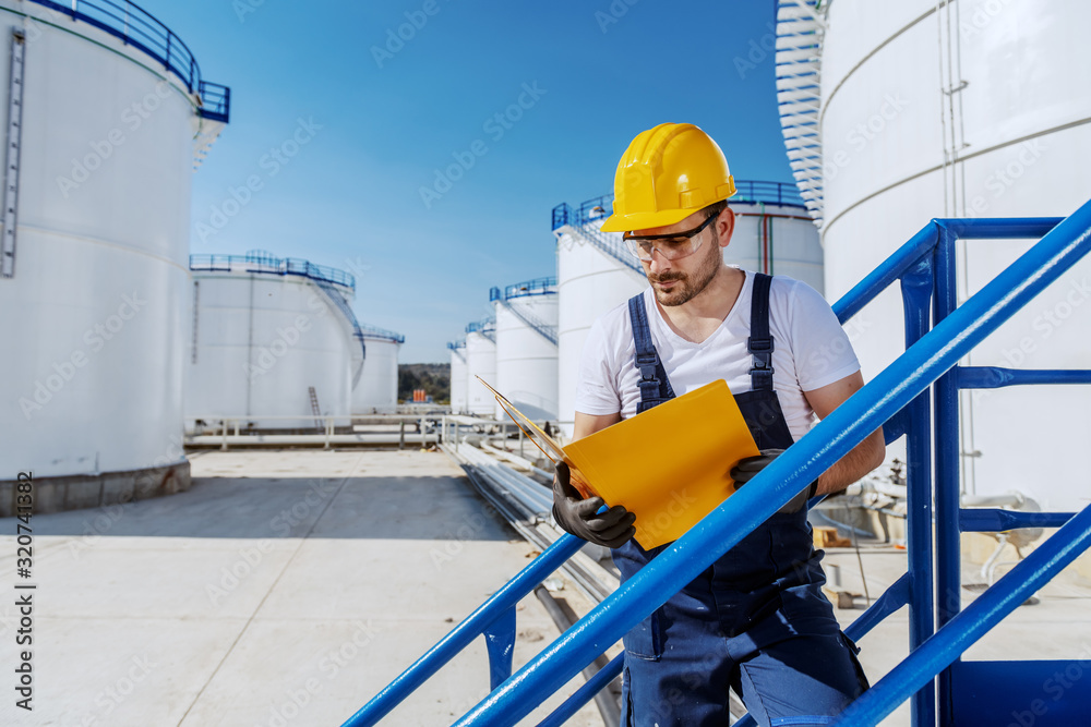 Hardworking handsome caucasian worker in overall and with helmet on head standing on the stairs and looking at folder with documents. Oil production. Hardworking handsome caucasian worker in overall and with helmet on head standing on the stairs and looking at folder with documents. Oil production.