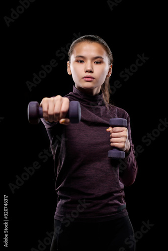 Young active female with dumbbells stretching arm forwards while exercising against black background in isolation