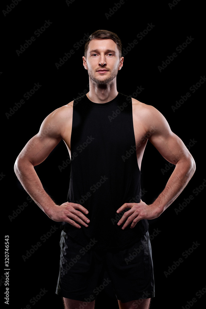 Fototapeta premium Young muscular athlete in black vest and shorts keeping hands on waist while standing in front of camera in isolation