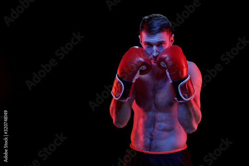 Young muscular boxer in boxing gloves standing in front of camera ready to fight his rival over black background