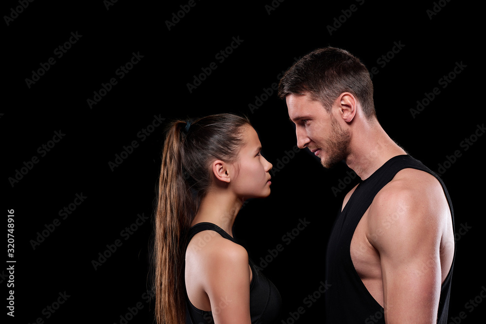 Young fit woman and man in activewear looking at one another while standing in front of camera on black background Young fit woman and man in activewear looking at one another while standing in front of camera on black background