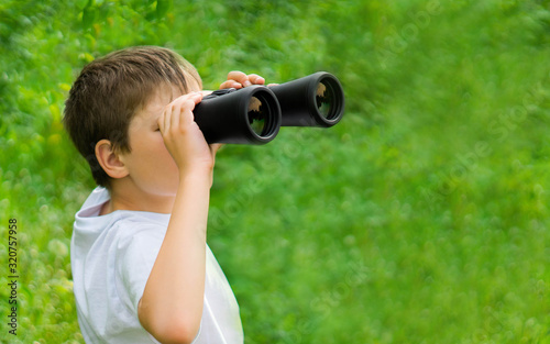 banner size. curious child boy in white t-shirt looking through binoculars in nature green landscape. Explore and discover concept. kid adventure. copy space for text.