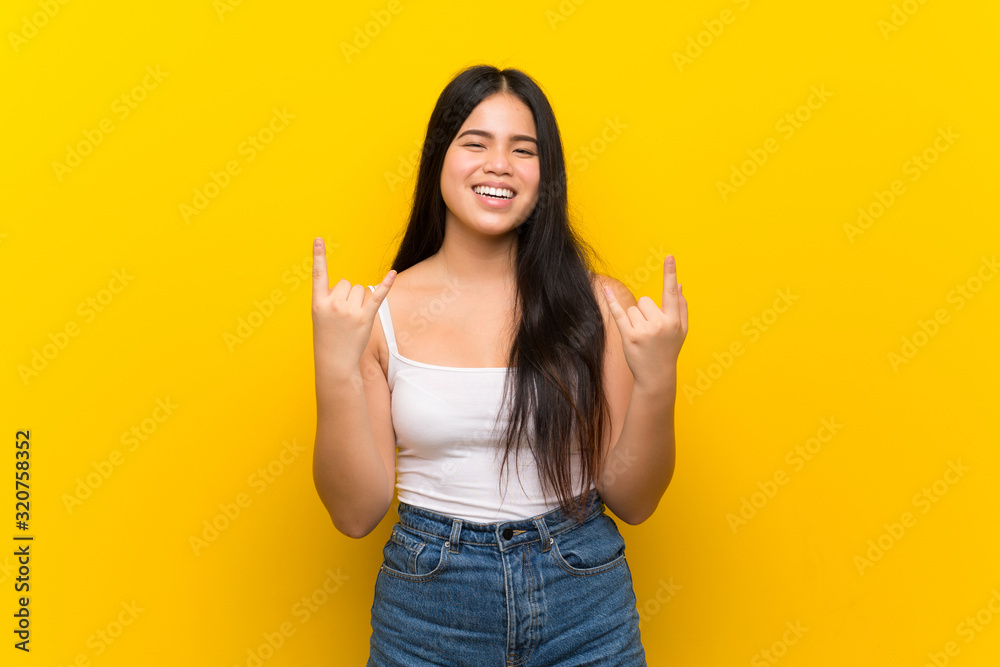 Young teenager Asian girl over isolated yellow background making rock gesture Young teenager Asian girl over isolated yellow background making rock gesture