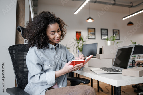 Cute African American woman makes notes with a pen in a notebook, putting off work and basic duties, taking a break for herself and time for rest. Creative office and creative work concept.