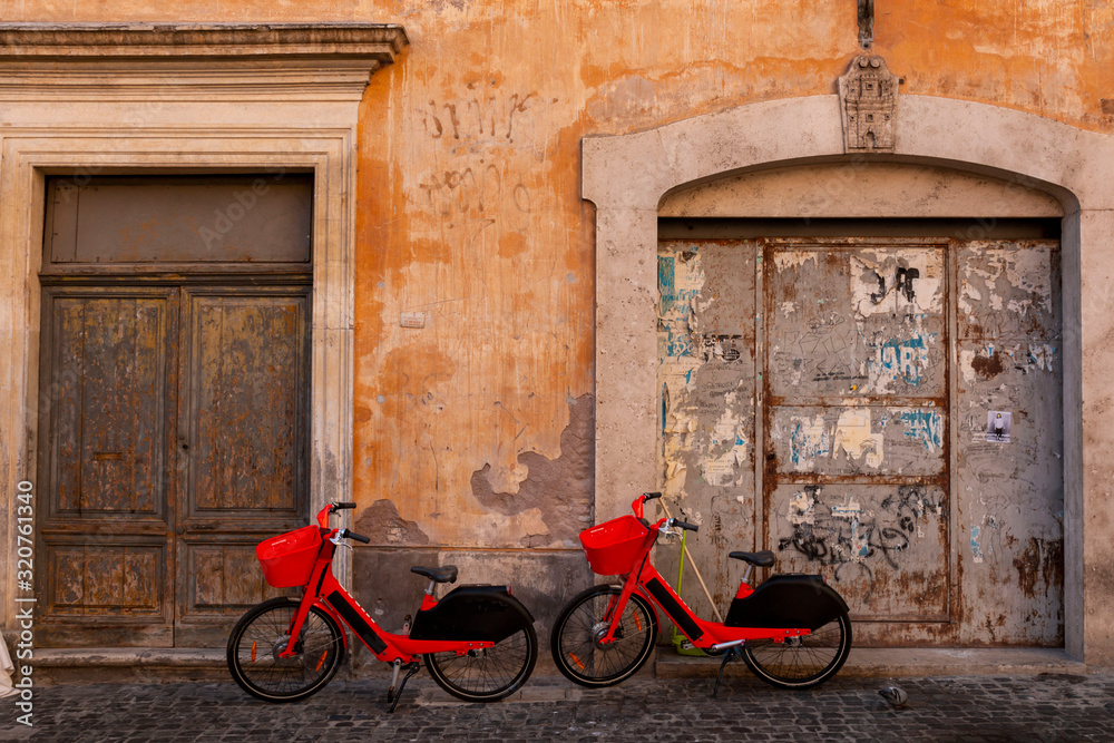 Red pedal-assist bikes parked by an old wall. Electric Bicycle sharing system rentals with basket on a sidewalk in street of Rome. Red pedal-assist bikes parked by an old wall. Electric Bicycle sharing system rentals with basket on a sidewalk in street of Rome.