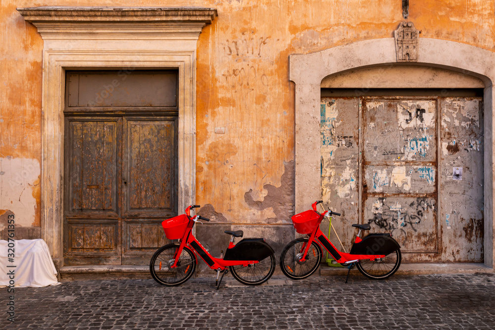 Red pedal-assist bikes parked by an old wall. Electric Bicycle sharing system rentals with basket on a sidewalk in street of Rome.