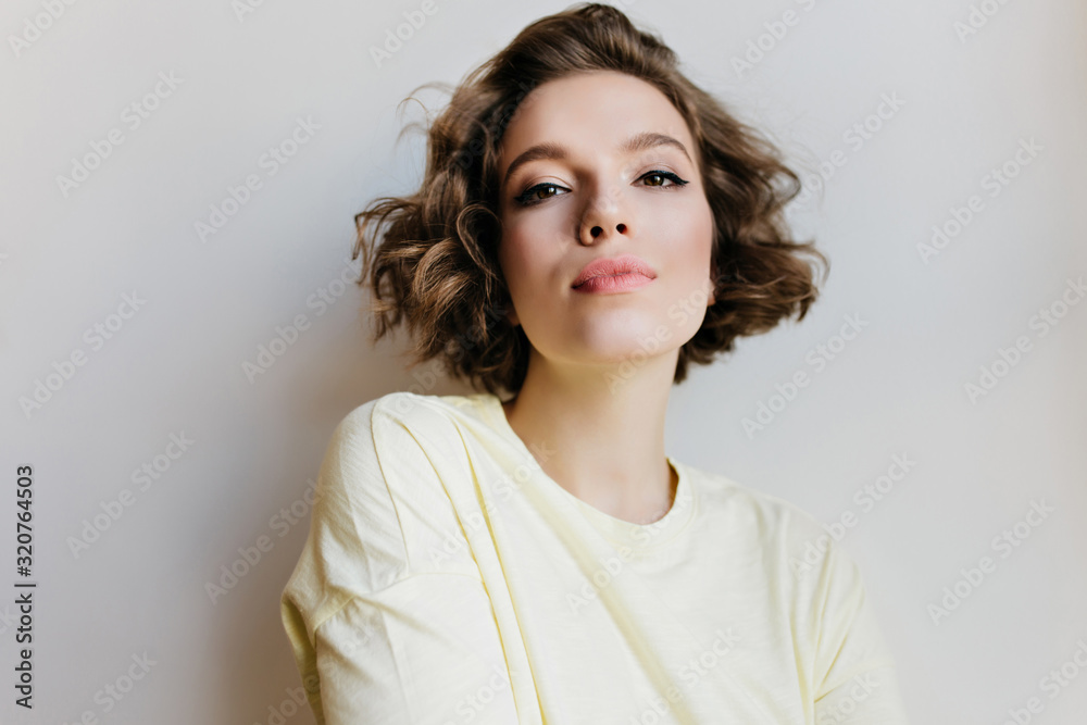 Pretty girl with casual makeup posing with interest on light background. Lovable short-haired lady with dark eyes standing in studio with serious face expression. Pretty girl with casual makeup posing with interest on light background. Lovable short-haired lady with dark eyes standing in studio with serious face expression.