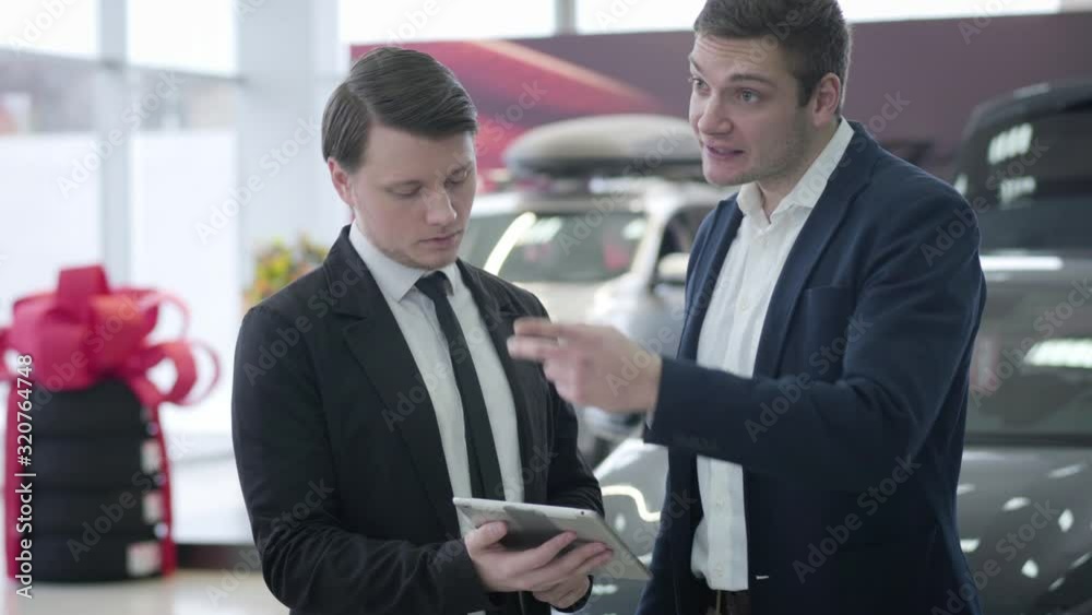 Portrait of male Caucasian car dealer and client using tablet and looking around in car showroom. Two men in business suits discussing purchase of automobile in car dealership. Business, lifestyle. Portrait of male Caucasian car dealer and client using tablet and looking around in car showroom. Two men in business suits discussing purchase of automobile in car dealership. Business, lifestyle.