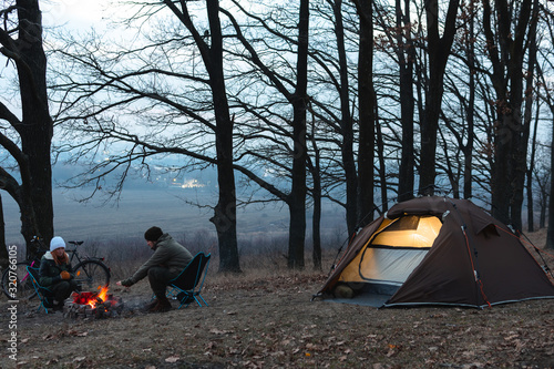A couple of people, man and woman are sitting on light chairs by the fire. Cold autumn evening in the forest. The concept of an active lifestyle, travel, vacation. Camping tent light. Bonfire