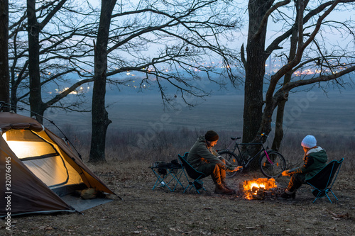 A couple of people, man and woman are sitting on light chairs by the fire. Cold autumn night in the forest. The concept of an active lifestyle, travel, vacation. Camping tent light. Bonfire