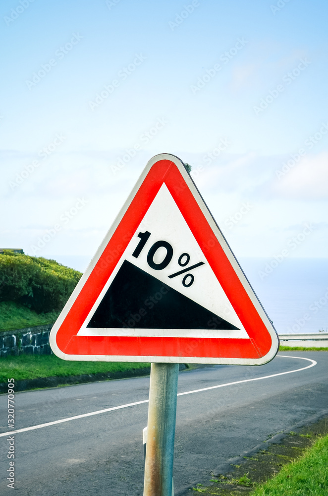 Red triangle road sign indicating a steep 10 percent downhill gradient in the road ahead. Empty road surrounded by green grass in the background. Traffic signs. Vertical photo