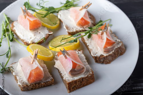Sandwiches with salmon on dark bread, spread with cheese sauce. The meat is wrapped in a slice of daikon and fixed with a decorative clothespin. On a wooden background.