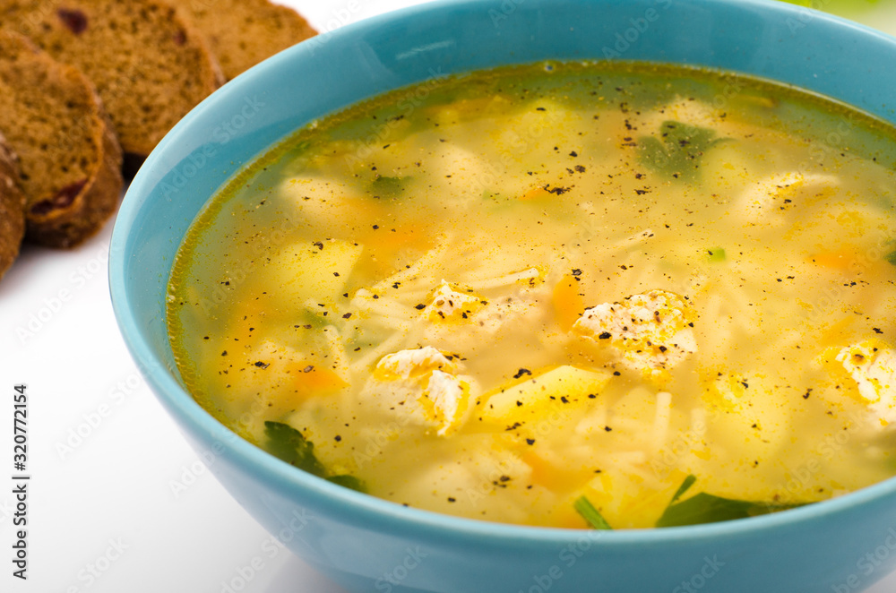 Chicken soup with vegetables in a blue plate on a white table, near a spoon and bread, closeup