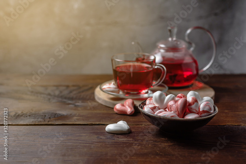 Beautiful romantic breakfast with red tea Hibiscus and meringue cookies in the shape of hearts. Glass tea-set stand on a dark wooden table with a concrete background. Sunny still life with copy space.