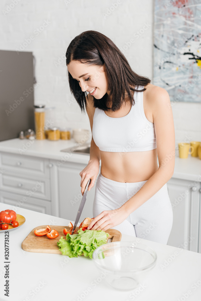 Smiling young woman cooking fresh salad with organic vegetables on kitchen table