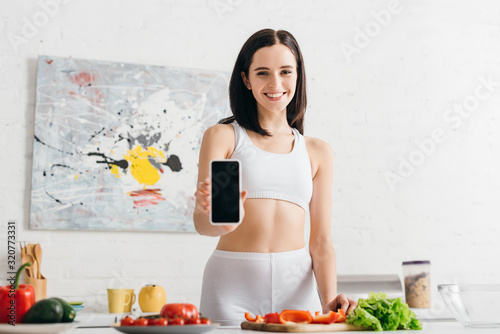 Beautiful sportswoman showing smartphone and smiling at camera while cooking fresh salad in kitchen