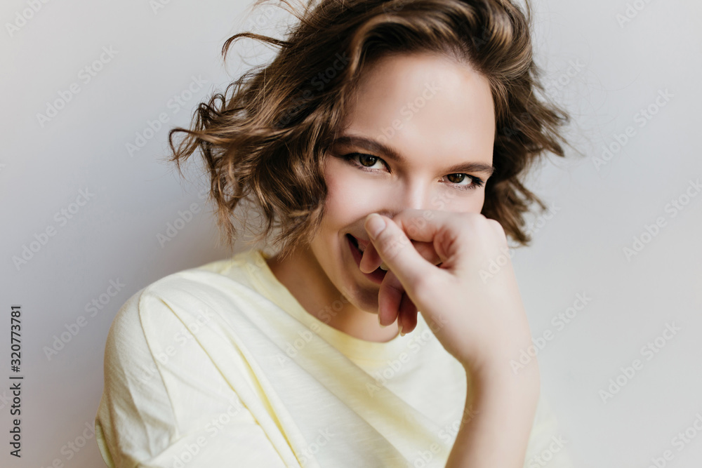 Close-up photo of charming curly lady covering face and laughing. Indoor portrait of carefree girl posing with shy smile on light background. Close-up photo of charming curly lady covering face and laughing. Indoor portrait of carefree girl posing with shy smile on light background.