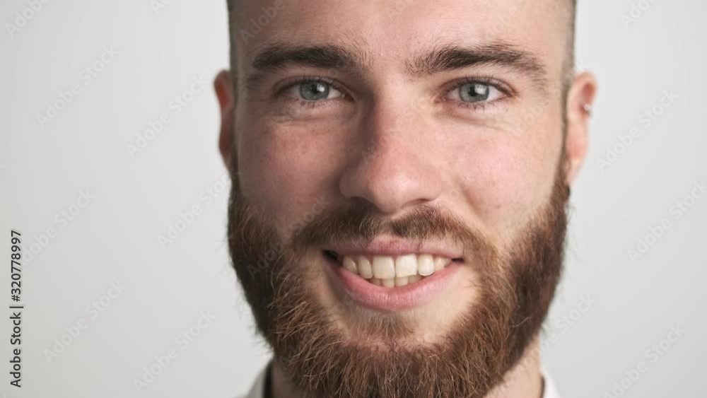 A close-up view of handsome bearded man raises his eyes to the camera with a smile isolated over white wall background A close-up view of handsome bearded man raises his eyes to the camera with a smile isolated over white wall background