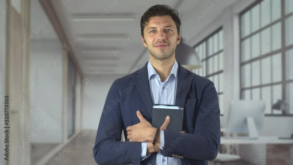 Portrait of handsome attractive young businessman in suit holding financial documents looking confidently at camera in the office. Happy young smart man. Shot in 6k. Portrait of handsome attractive young businessman in suit holding financial documents looking confidently at camera in the office. Happy young smart man. Shot in 6k.