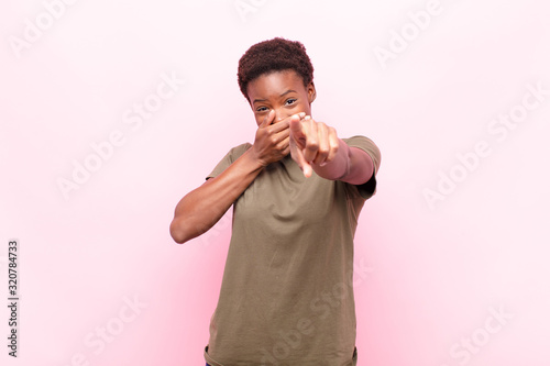 young pretty black womanlaughing at you, pointing to camera and making fun of or mocking you against pink wall photo