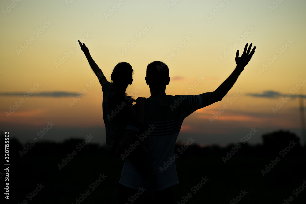 Silhouettes of father and daughter on his shoulders with hands up having fun, against sunset sky. Parenthood, family activities, vacation, support and love themes