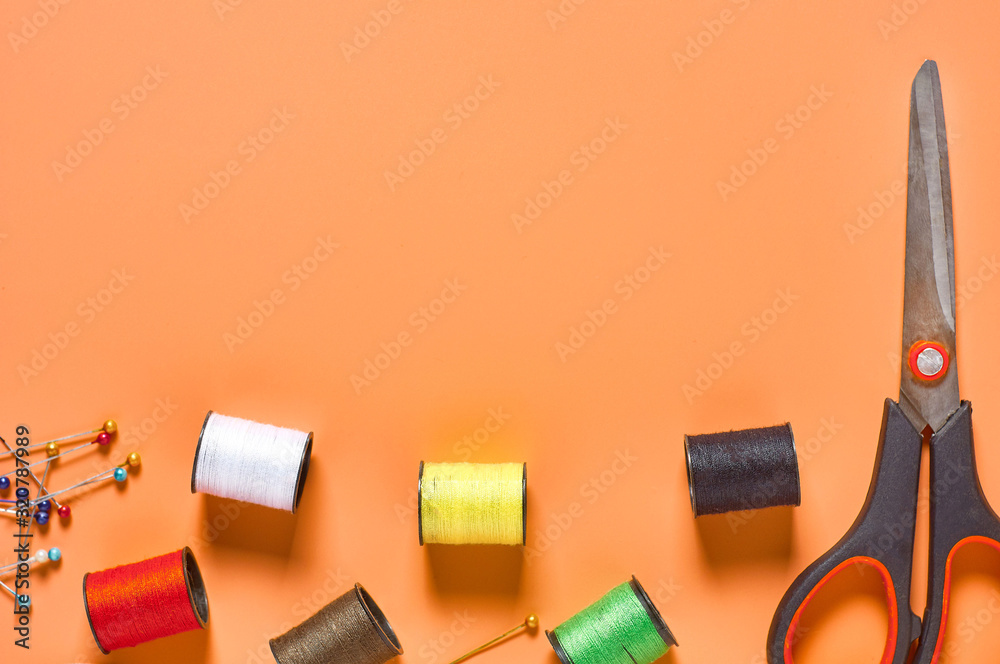 Scattered sewing accessories needles, scissors and spools with colorful threads on orange tailors desk in workshop. Space for text. Top view Scattered sewing accessories needles, scissors and spools with colorful threads on orange tailors desk in workshop. Space for text. Top view