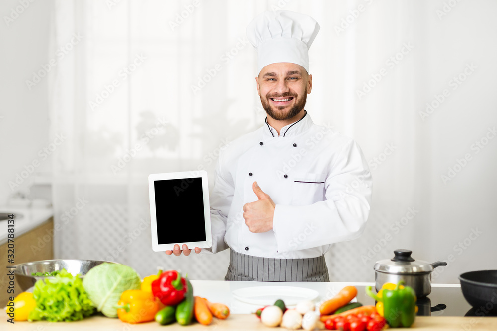 Chef Man Showing Tablet Screen Gesturing Thumbs-Up Standing In Kitchen Chef Man Showing Tablet Screen Gesturing Thumbs-Up Standing In Kitchen
