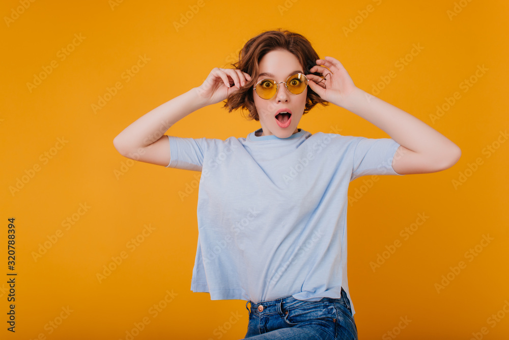 Studio portrait of gorgeous woman in light-blue t-shirt expressing amazement. Indoor photo of emotional lady with wavy hairstyle looking through yellow sunglasses. Studio portrait of gorgeous woman in light-blue t-shirt expressing amazement. Indoor photo of emotional lady with wavy hairstyle looking through yellow sunglasses.