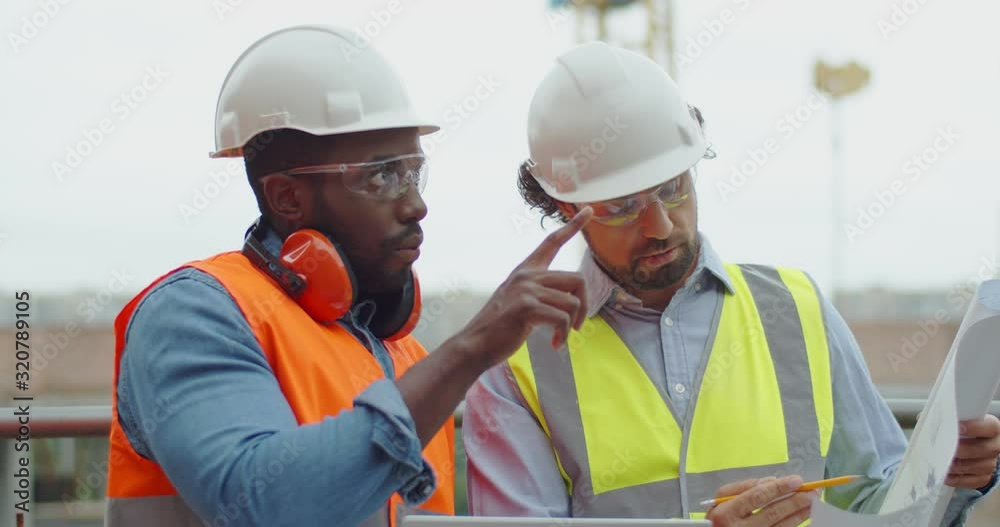 Close up of the two mixed-races male constructors in goggles and hardhats having a talk and discussion about building cunstruction plan. Outside. Close up of the two mixed-races male constructors in goggles and hardhats having a talk and discussion about building cunstruction plan. Outside.