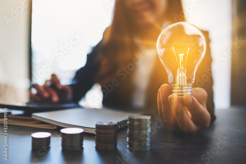 Businesswoman holding a lightbulb with coins stack on table, saving energy and money concept