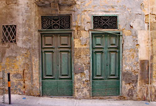 Two derelict green doors with ornate grills above them in a crumling limestone wall in Sliema, Malta
