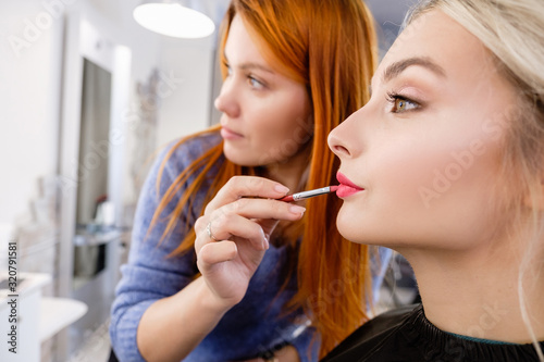 Hand of a professional make-up artist s master dyes the red lips of a young blonde client girl while creating makeup for a photo shoot in beauty room or luxury spa salon. Fashion shooting concept