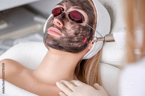 Close-up of cosmetologist's hand making hardware carbon peeling by young pretty customer woman in safety glasses headdress lying on couch in beauty salon. Facial cleaning concept. Cosmetology Services photo