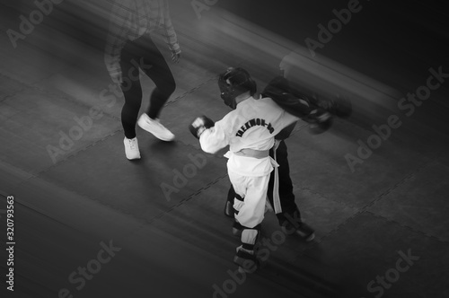 Kids in traditional kimano, hard hats and gloves. Sports duel. Black and white. For atmospheric effects, added motion blur effect and film noise. Text: Taekwondo is the name for martial art.